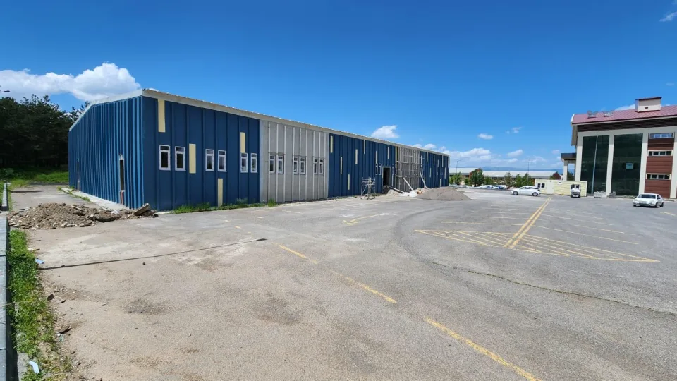 Mavi ve gri renkli geniş bir endüstriyel bina ile yanında çok katlı bir idari binanın bulunduğu açık otopark alanı.